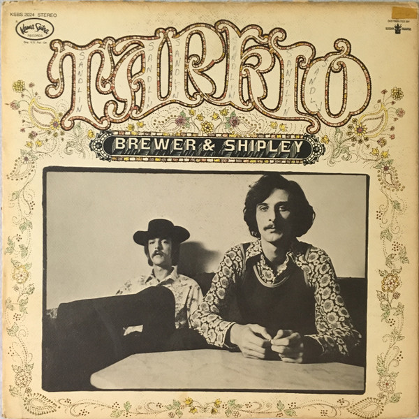 Album cover for Brewer & Shipley's Tarkio, showing the duo seated at a table in a black-and-white photo, framed by ornate lettering and a paisley border.