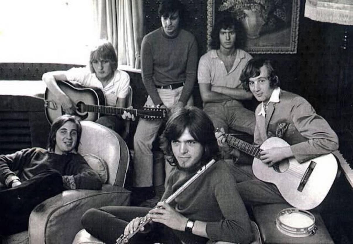Genesis band history begins in 1969: black and white photo of six young men in a sitting room with acoustic guitars and a flute, Genesis in the summer of 1969